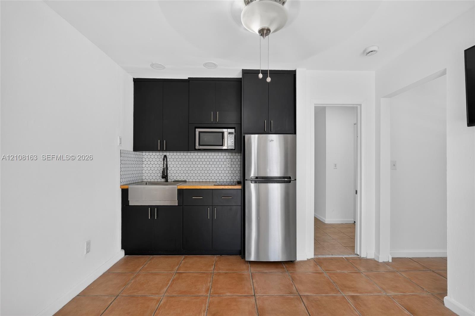 Kitchenette featuring black cabinetry, stainless steel refrigerator, apron-front sink, hexagon tile backsplash, and tile flooring
