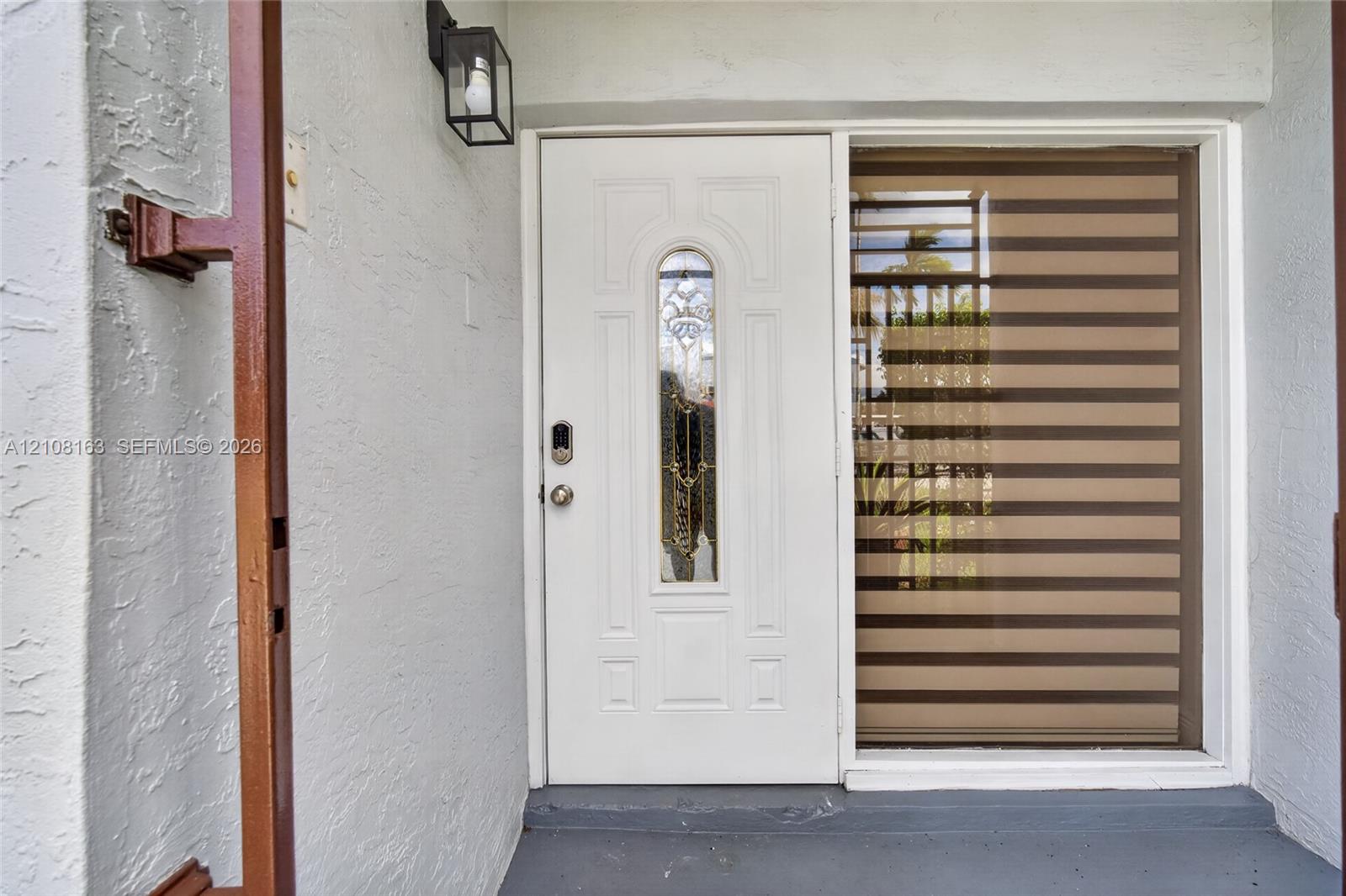 Entryway featuring a white exterior door with a decorative glass insert, integrated smart lock, metal wall-mounted lantern, and sidelight window with horizontal banded privacy shades