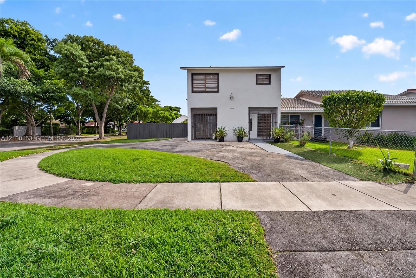 Two-story exterior with a light-toned stucco facade, paved driveway, concrete walkway, and chain-link perimeter fencing