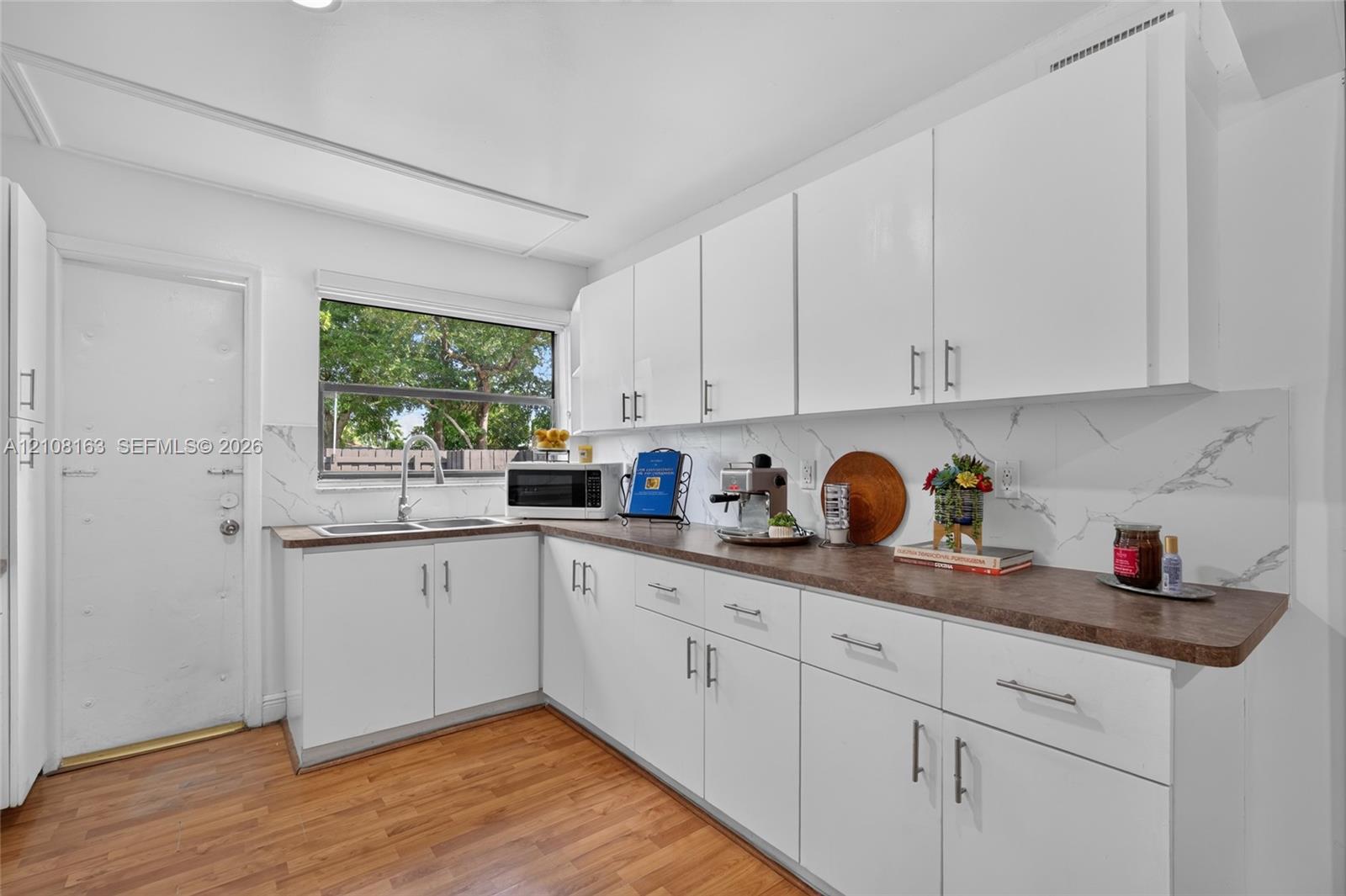 Kitchen featuring white cabinetry, wood-finish flooring, dark-toned countertops, and a dual-basin stainless steel sink with a stone-patterned backsplash