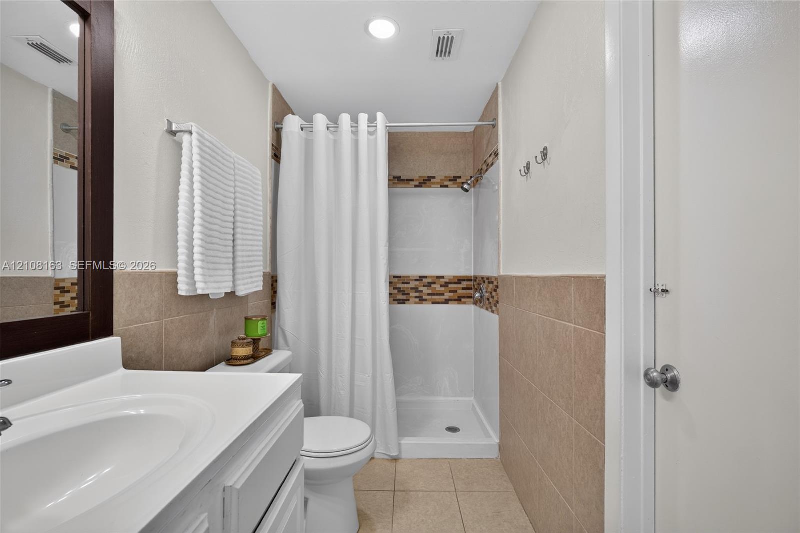 Bathroom featuring a white vanity with a porcelain sink, beige wall tiling, mosaic tile accent border, and a walk-in shower with white surround