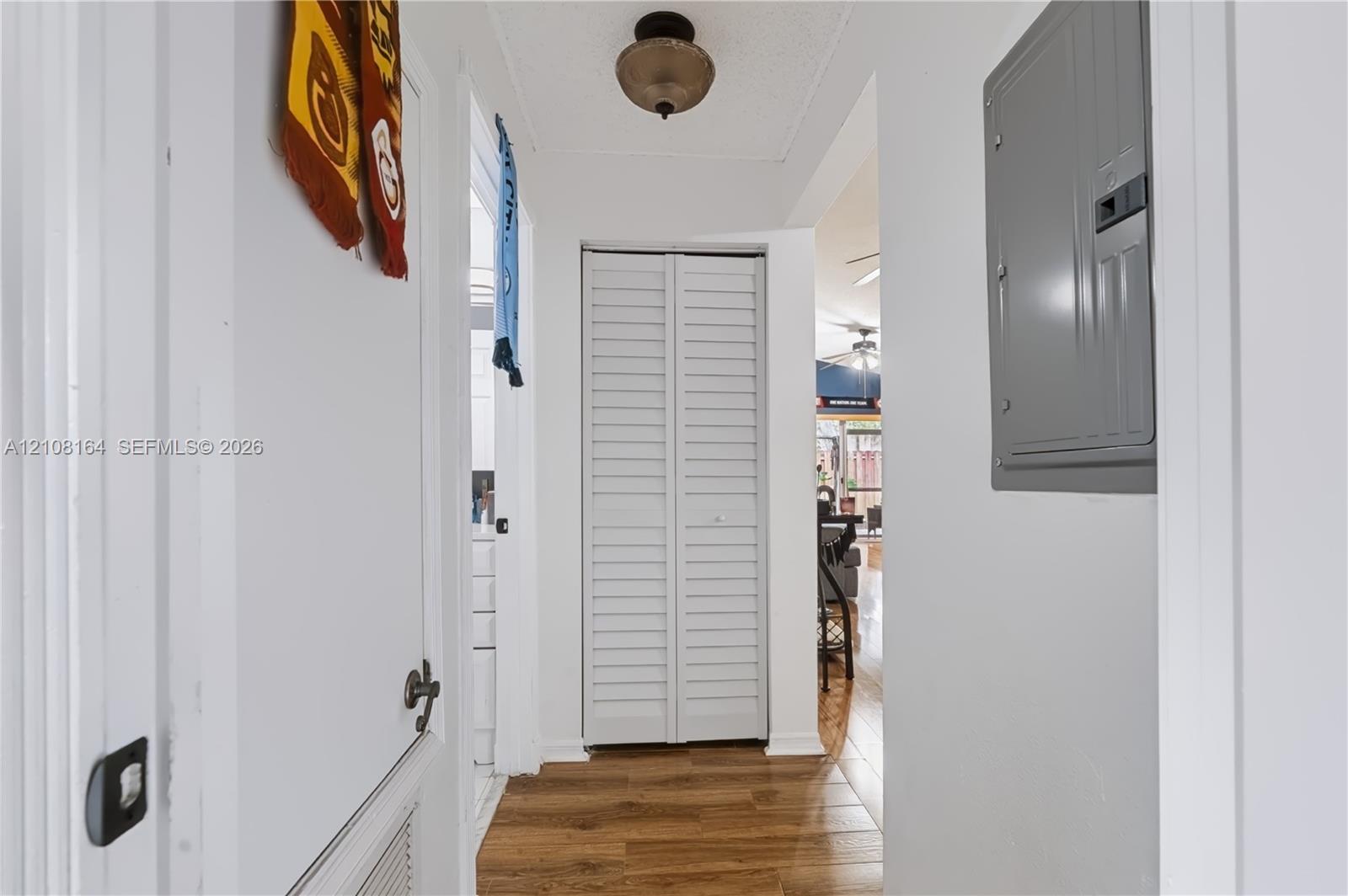 Hallway featuring wood-finish flooring, white louvered bifold closet doors, a neutral-tone flush-mount ceiling light fixture, and a wall-mounted electrical panel