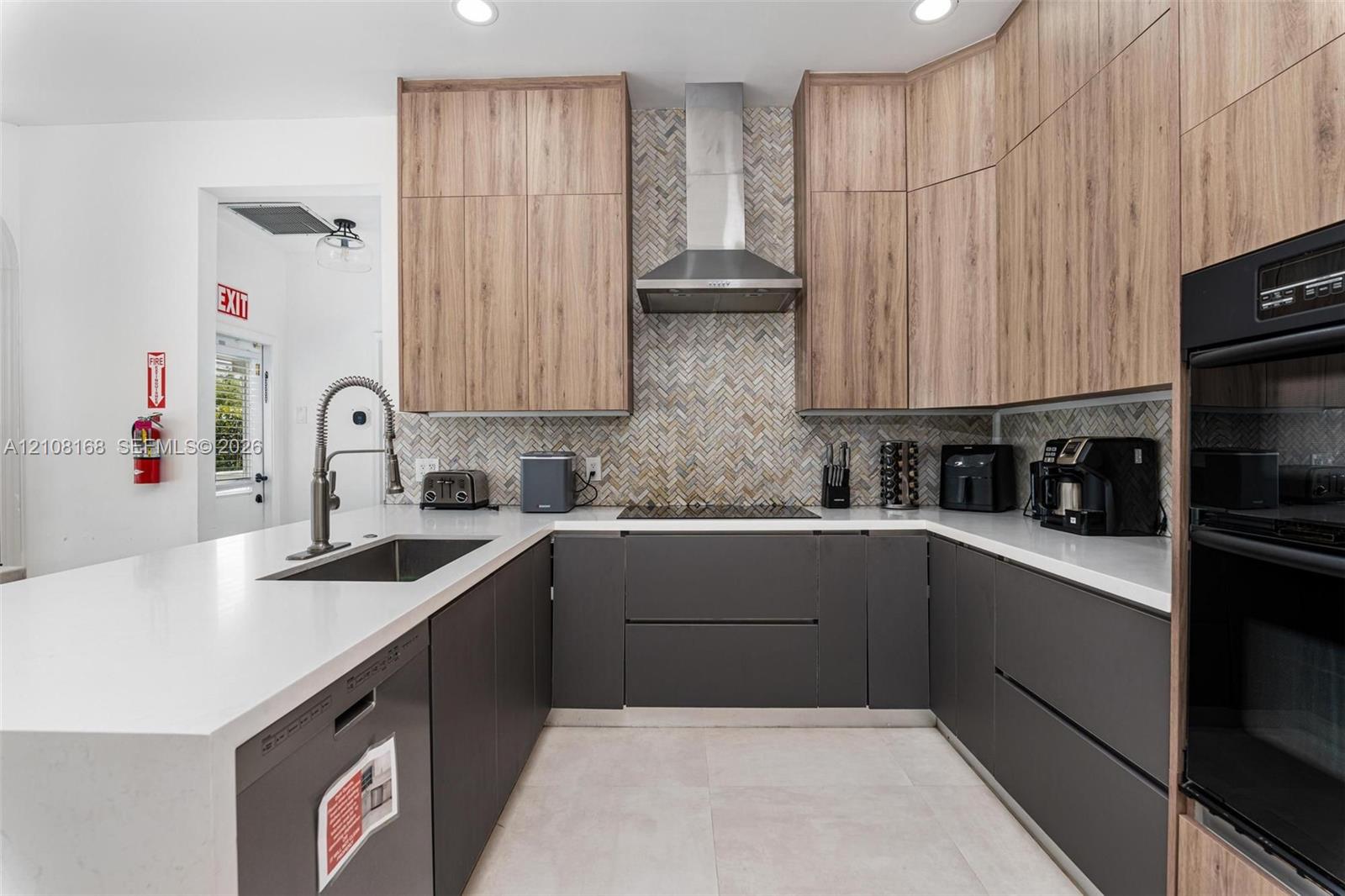 Kitchen featuring dual-tone cabinetry with wood-finish uppers, quartz countertops, herringbone-patterned tile backsplash, stainless steel range hood, and integrated wall oven