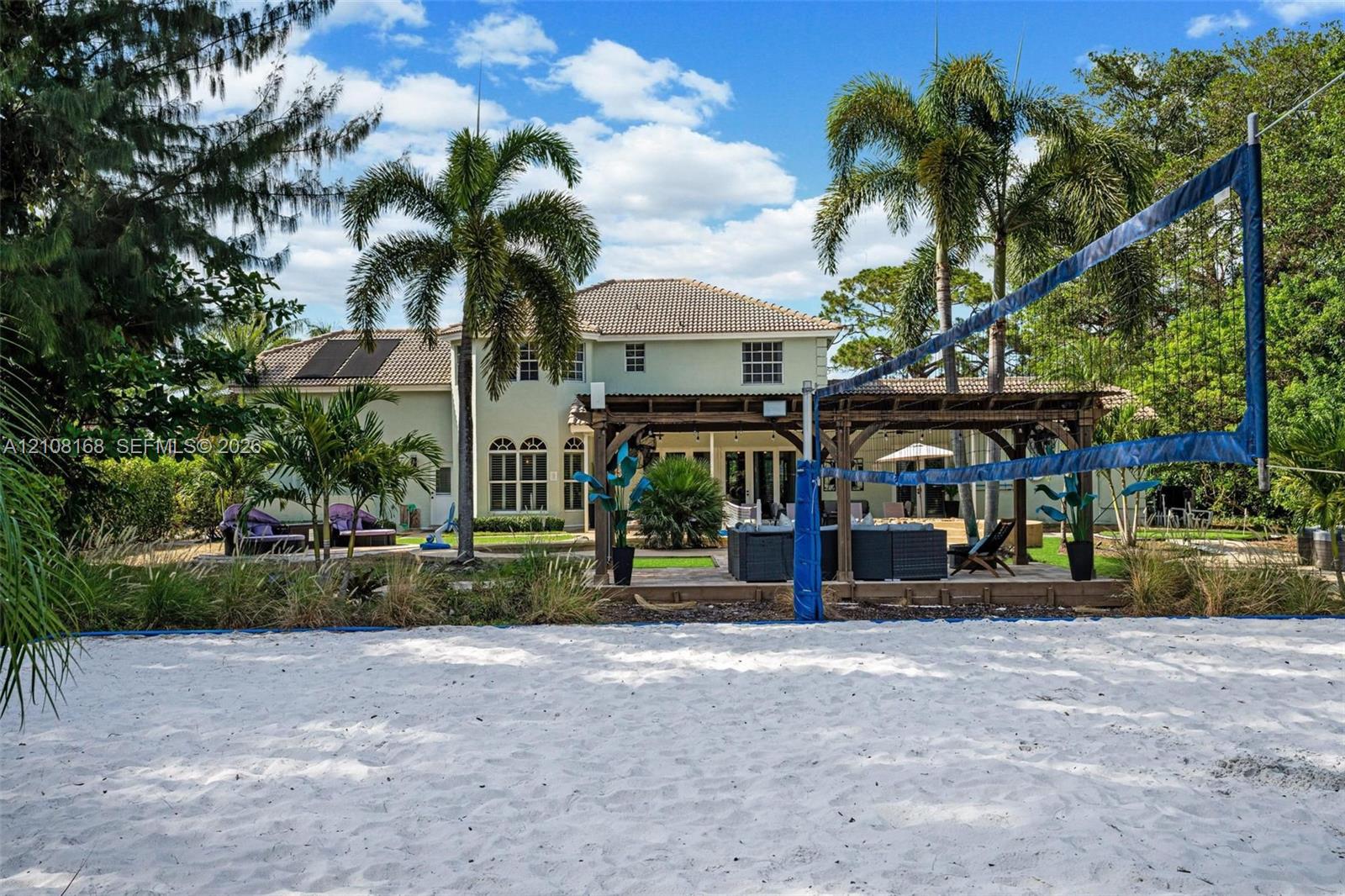 Sand volleyball court featuring a perimeter net and established palm trees, showcasing a two-story exterior with a covered pergola and masonry patio
