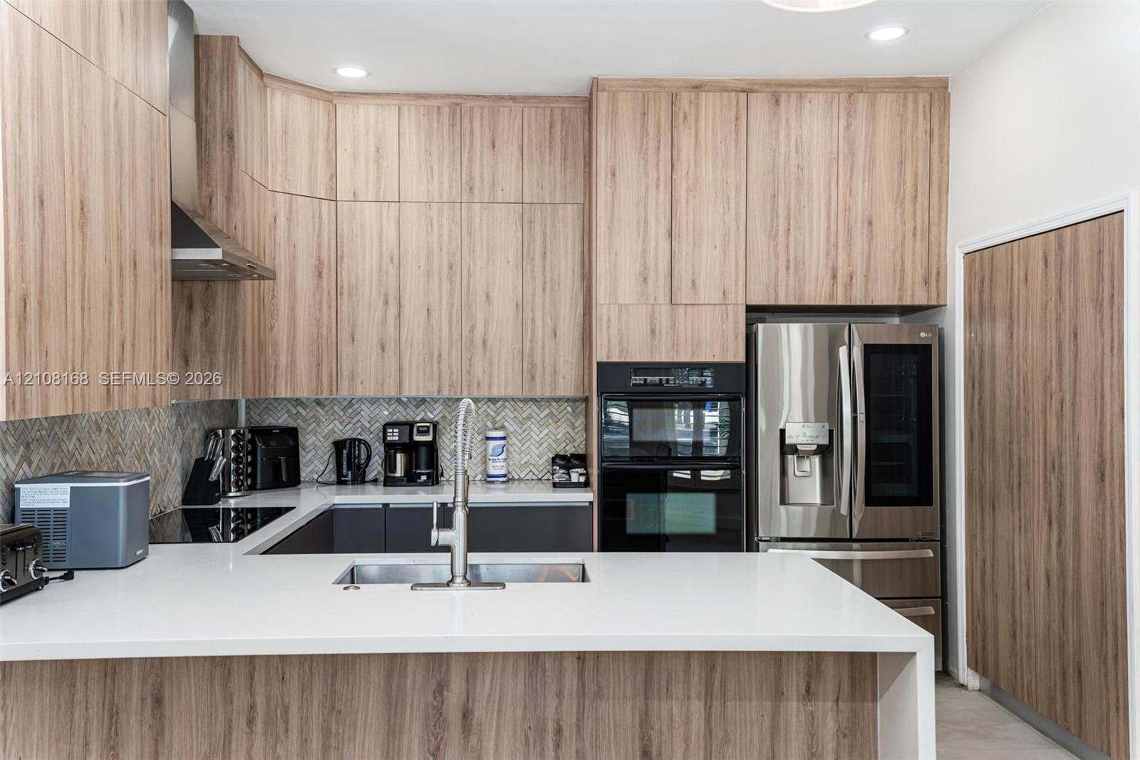 Kitchen featuring wood-finish cabinetry, white solid-surface countertops, a herringbone-patterned tile backsplash, a stainless steel undermount sink with an arched pull-down faucet, and built-in stainless steel appliances