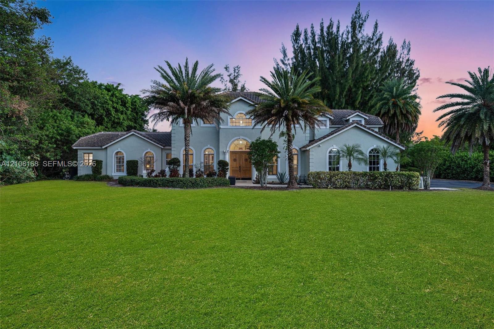 Multi-level residence featuring a stucco exterior, arched window detailing, double entry doors with glass insets, and expansive manicured grounds with mature palm trees