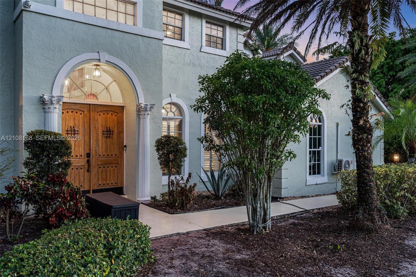 Stucco exterior featuring double arched wood front doors with decorative iron detailing, classical architectural columns, arched windows, and a paved walkway