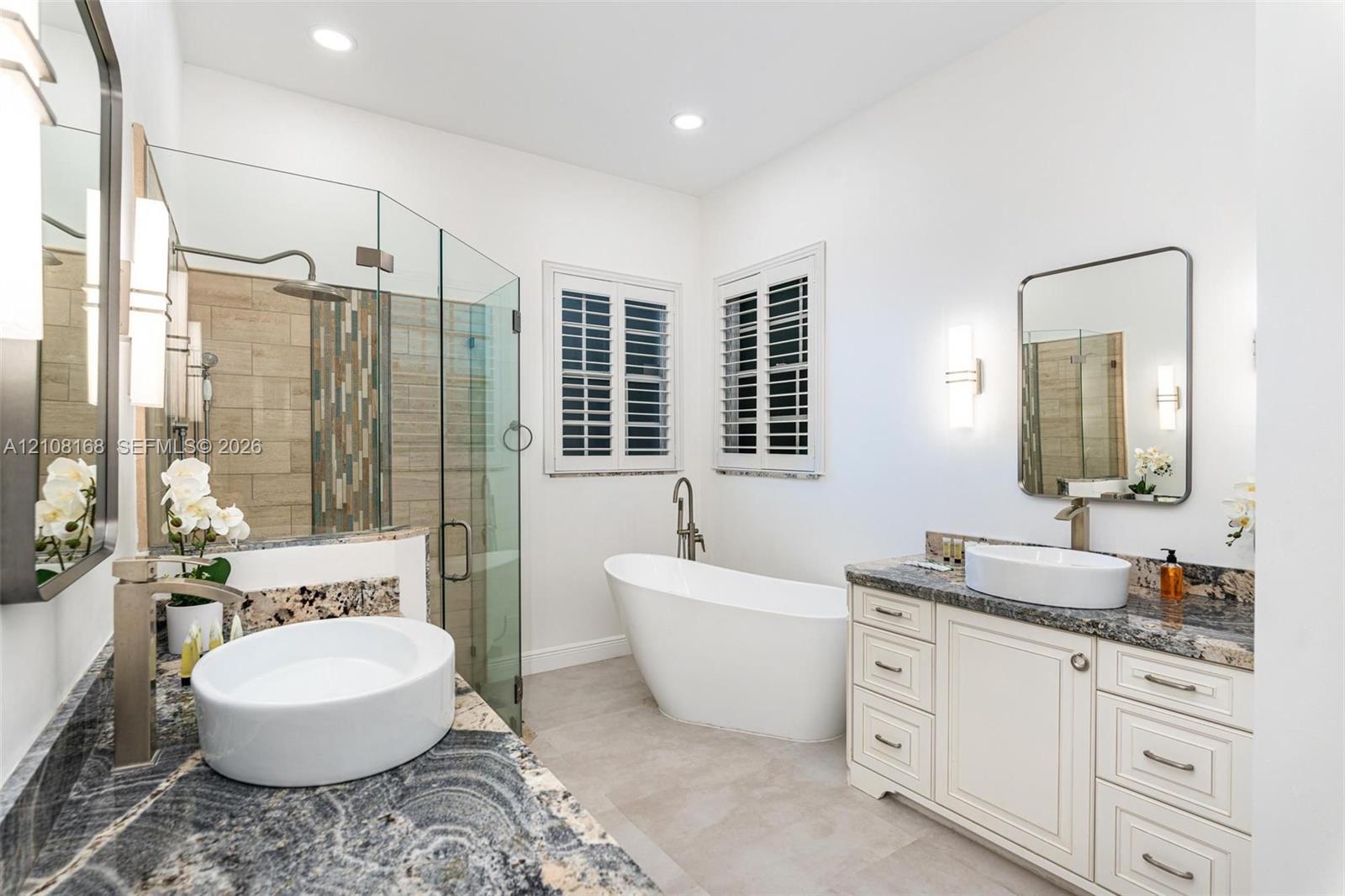 Dual vanities featuring stone countertops and vessel sinks, freestanding soaking tub, glass-enclosed shower with tiled walls, and plantation shutters