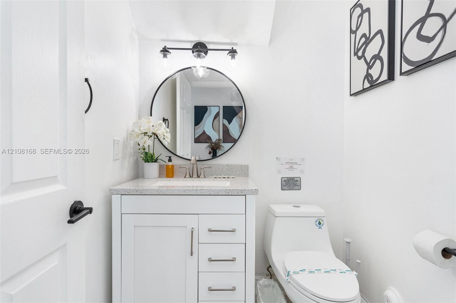 White vanity cabinet with a speckled countertop, integrated sink, circular wall-mounted mirror, three-light matte black fixture, and low-profile toilet