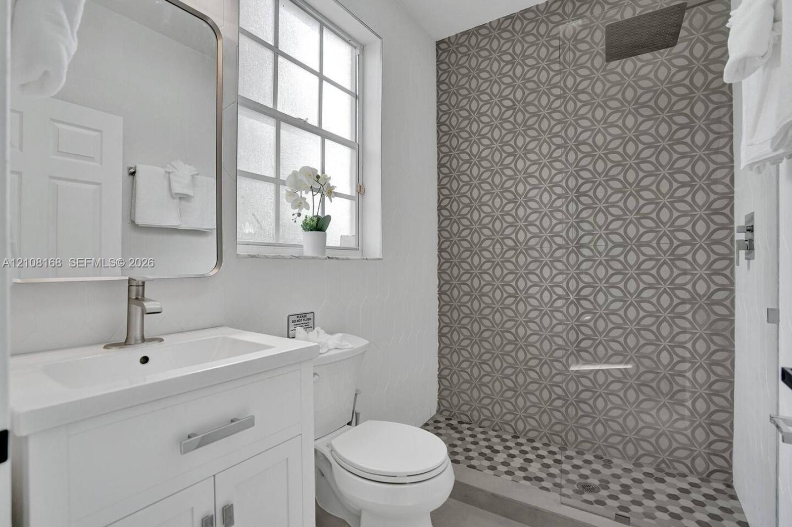 Bathroom featuring a white vanity cabinet with a rectangular basin, patterned geometric tilework in the walk-in shower, recessed ceiling-mounted shower head, glass shower panel, and a grid-pane window