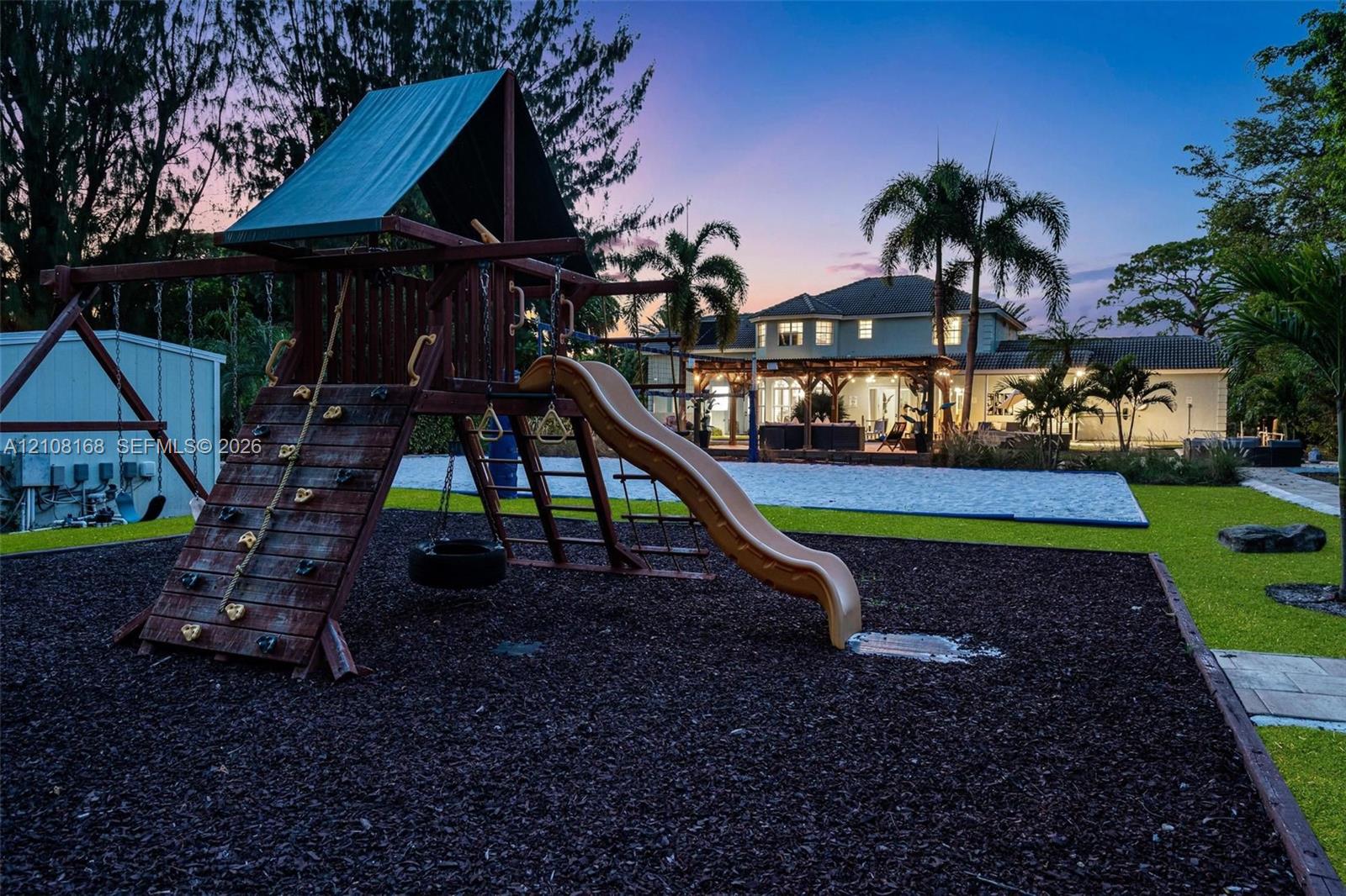 Expansive backyard featuring a built-in wooden play structure with a climbing wall and slide, manicured lawn, established palm trees, and a paved patio area