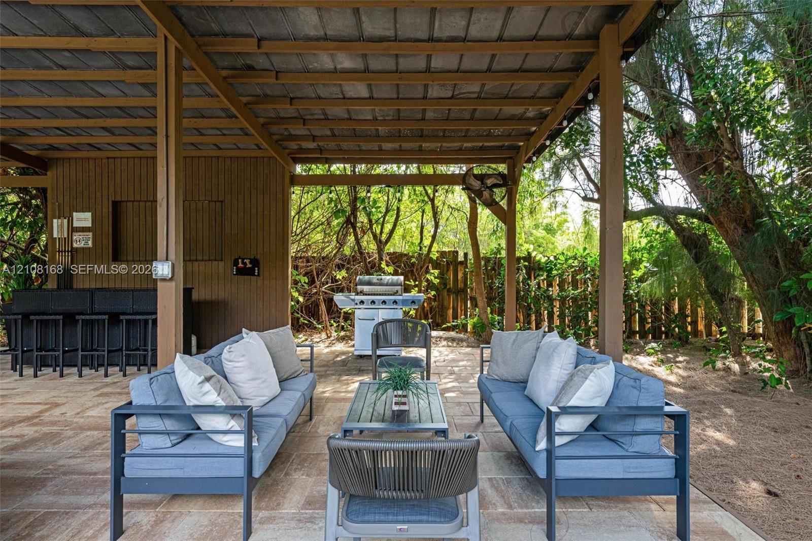 Covered outdoor patio featuring wood-paneled walls, a built-in bar, stone-paver flooring, and an attached ceiling fan