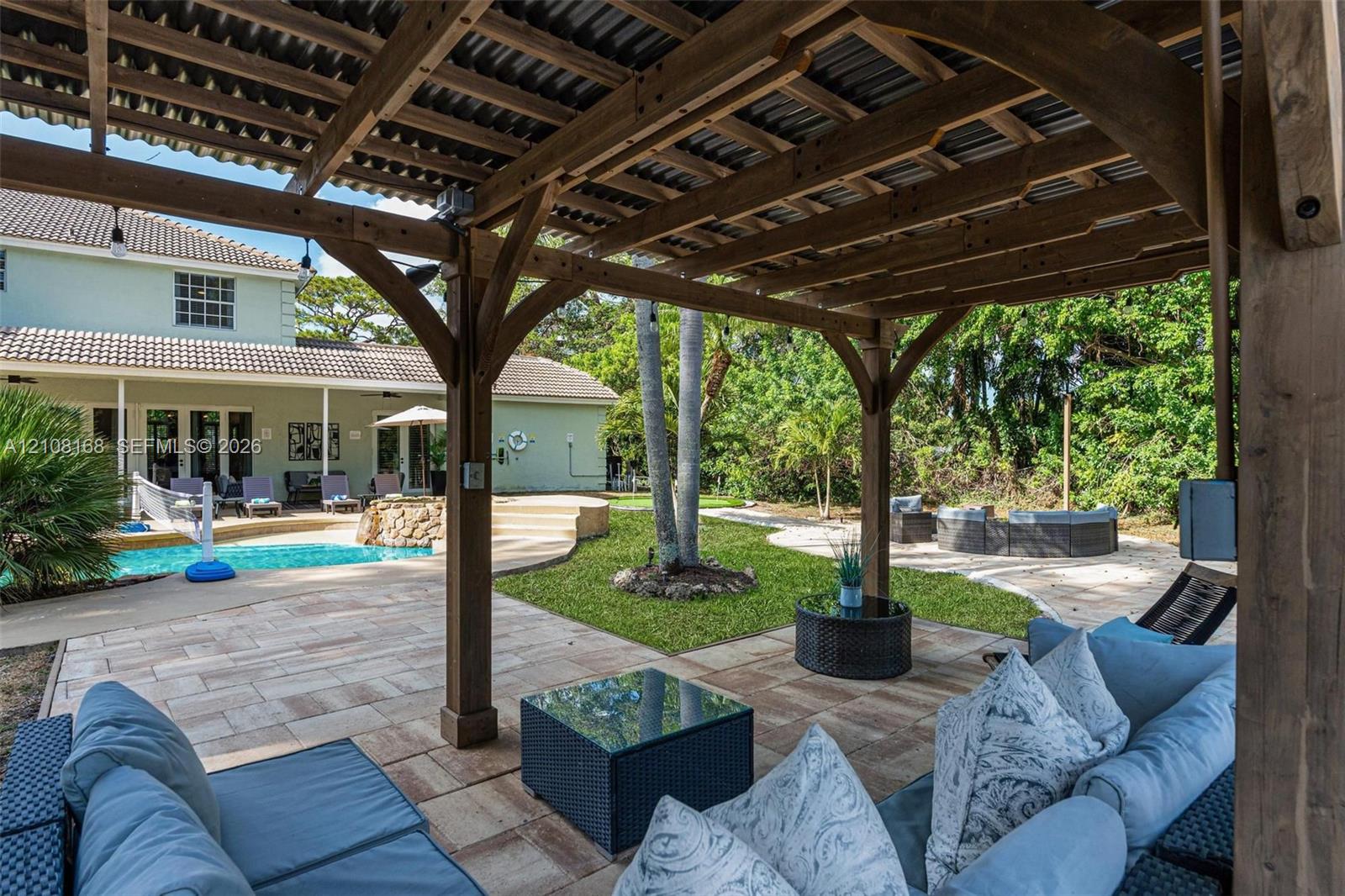 Covered wood-frame pergola with a corrugated roof, expansive stone-paver patio, swimming pool with a raised stone-faced spillway, and mature landscaping
