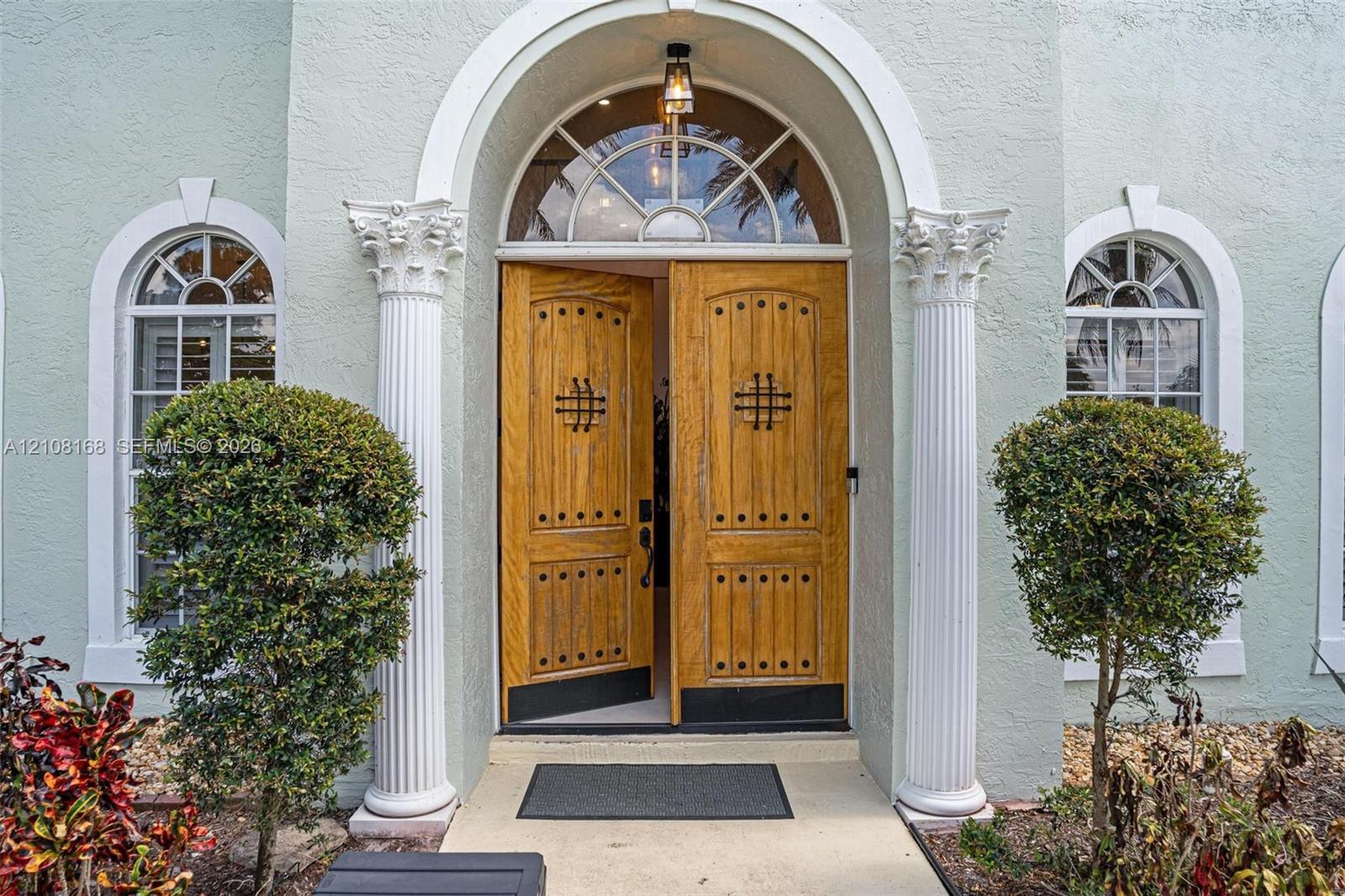 Entryway featuring dual wood-finish doors with decorative metal accents, an arched transom window, fluted columns with ornate capitals, and arched windows