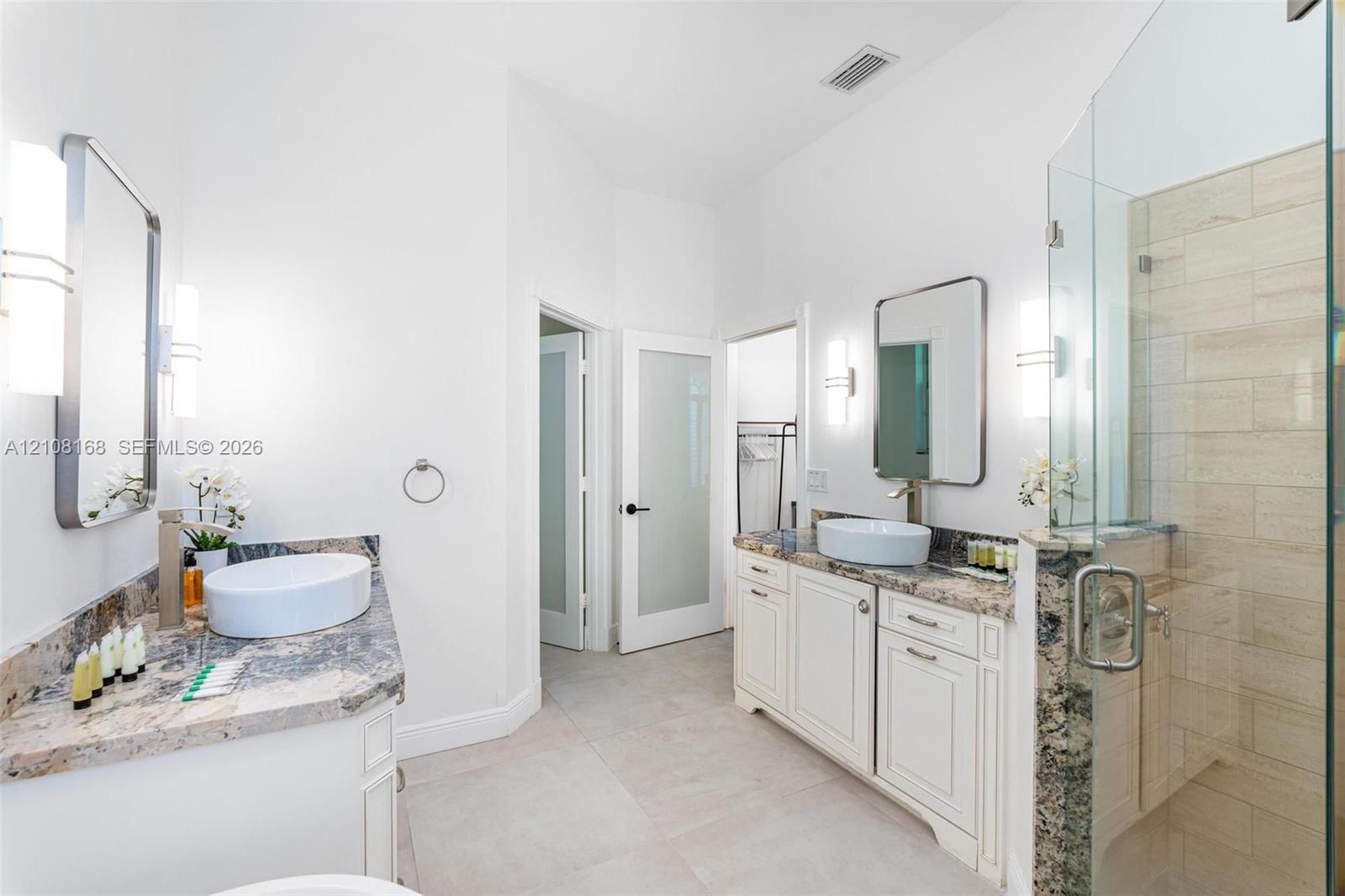 Dual vanity featuring stone countertops, vessel sinks, wall-mounted light fixtures, glass-enclosed shower with tile surround, and frosted glass door