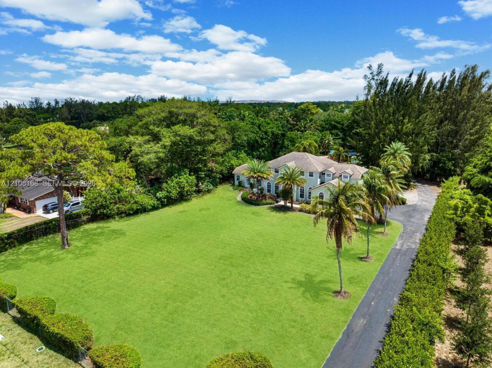 Expansive lot featuring a multi-story residence, extensive lawn area, mature palms, long paved driveway, and perimeter landscaping
