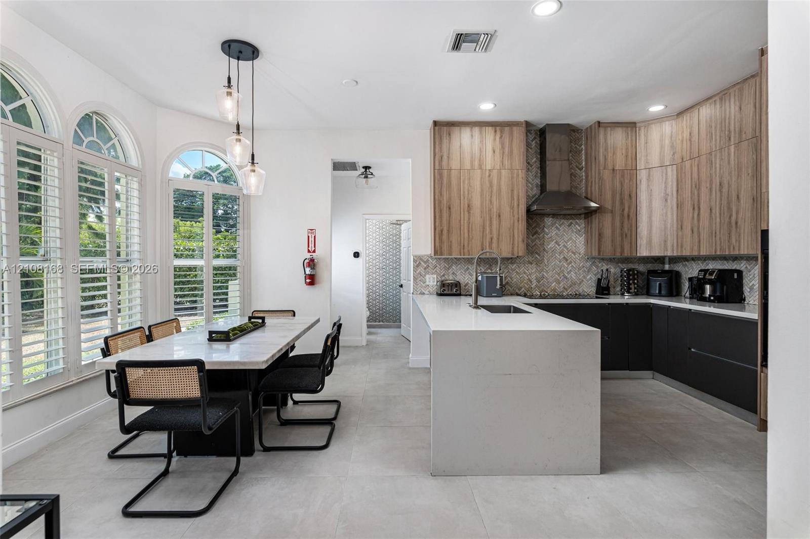 Kitchen featuring two-tone flat-panel cabinetry, a waterfall-edge center island, stainless steel range hood, herringbone tile backsplash, and arched windows with integrated shutters