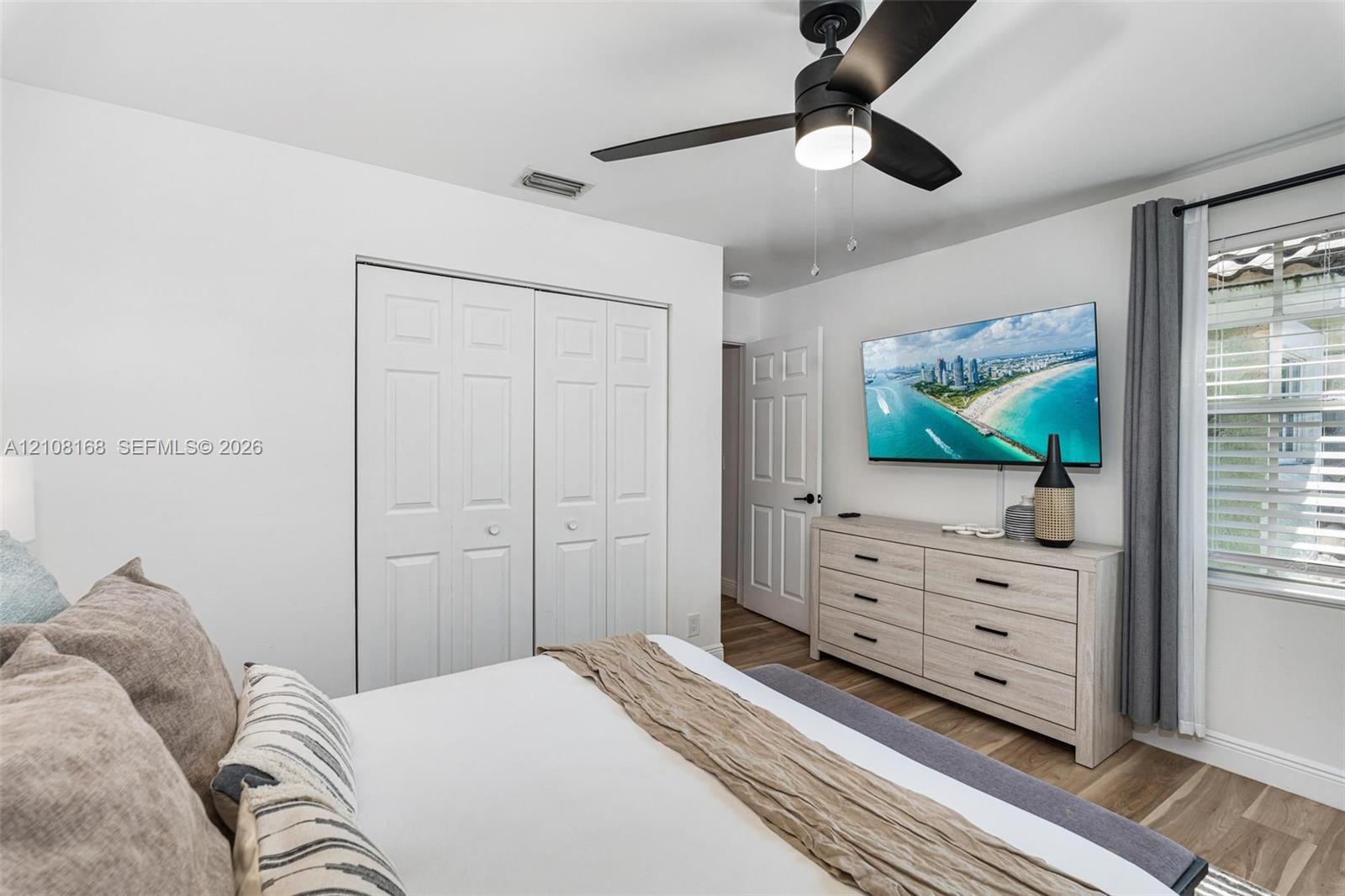 Room featuring wood-finish flooring, a ceiling fan with integrated lighting, dual-panel closet doors, and multi-pane window with horizontal blinds