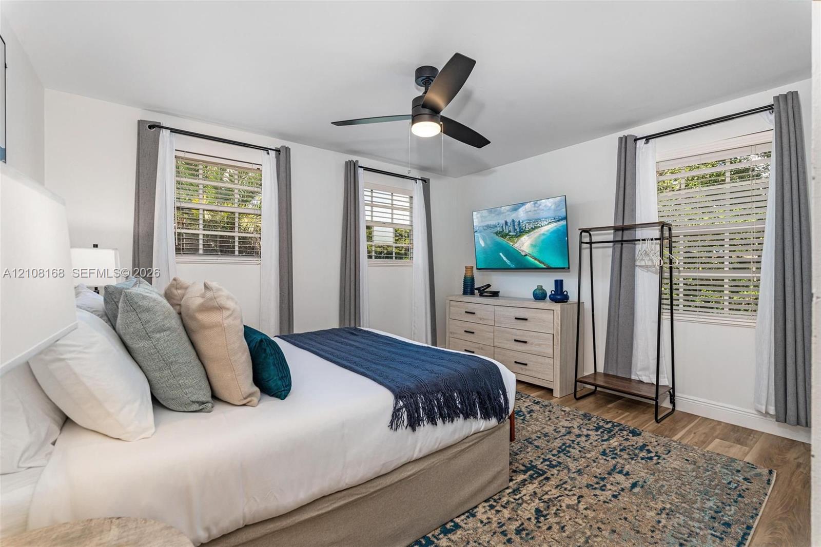 Bedroom featuring wood-finish flooring, three windows with plantation-style shutters, and a central ceiling fan with integrated lighting