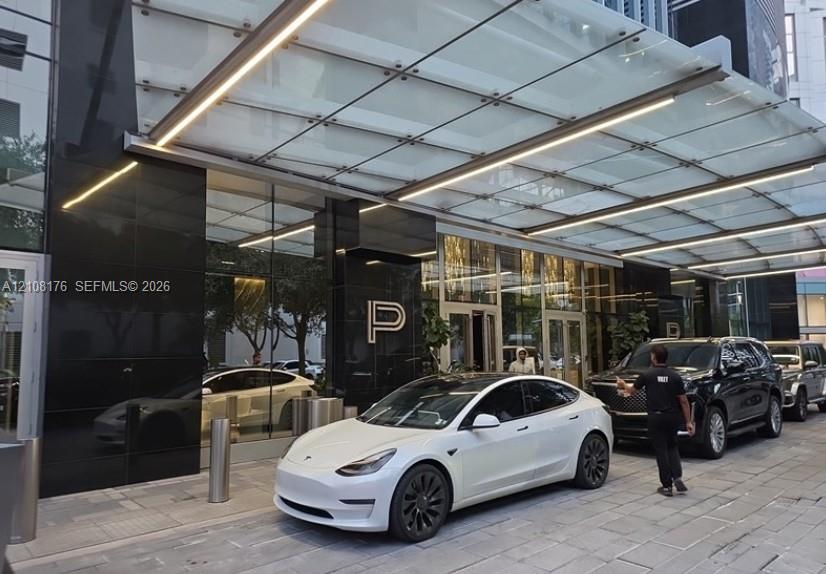 Covered porte-cochre featuring a glass canopy with integrated linear lighting, polished black stone facade, and stone-paved motor court