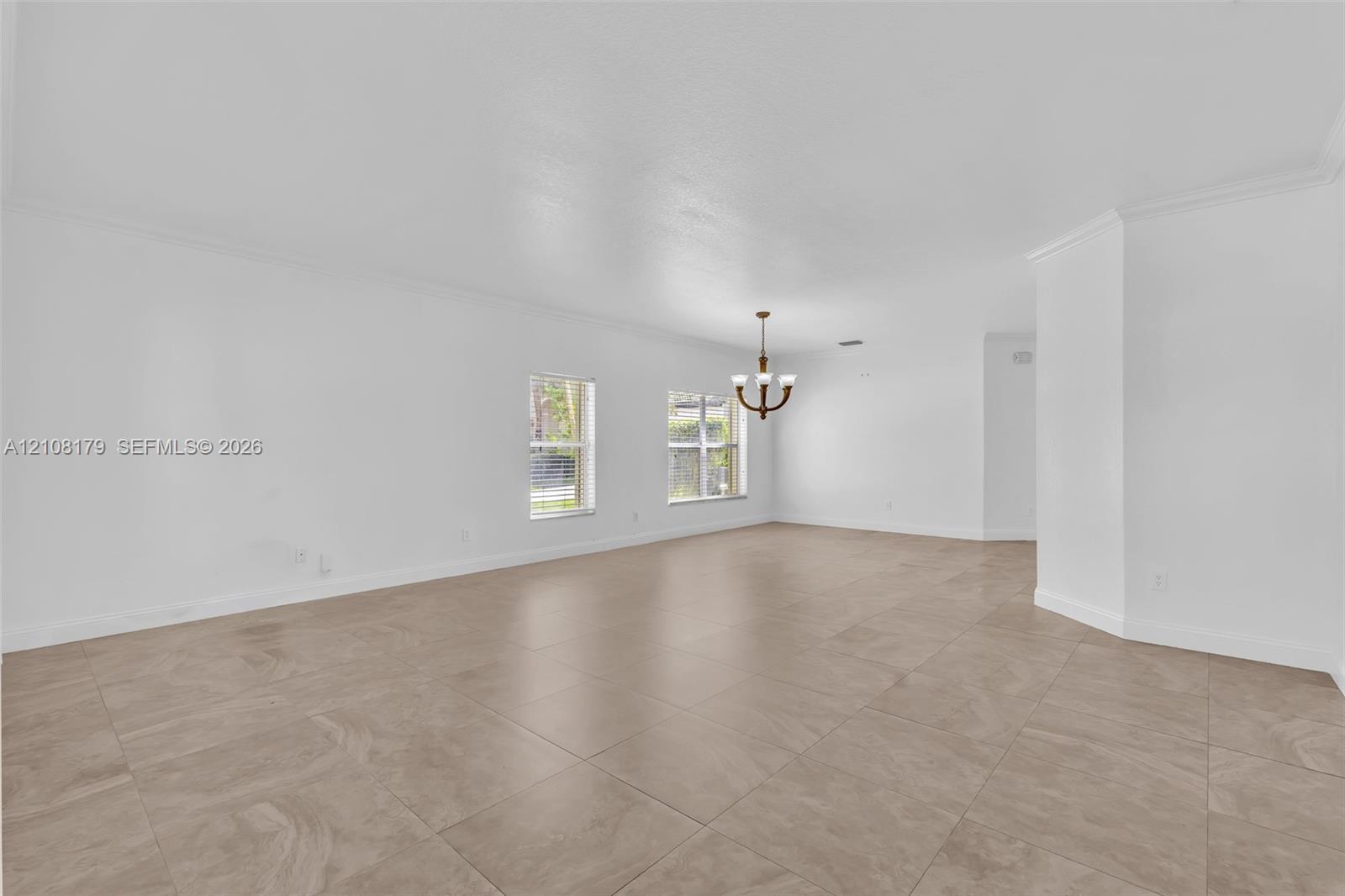 Open-concept interior featuring large-format tile flooring, recessed crown molding, dual windows, and a multi-arm chandelier