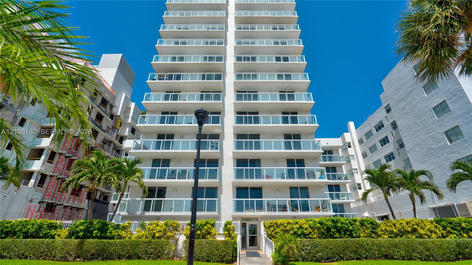 Multi-story high-rise exterior featuring stacked glass-railing balconies, floor-to-ceiling windows, white facade, and manicured landscaping