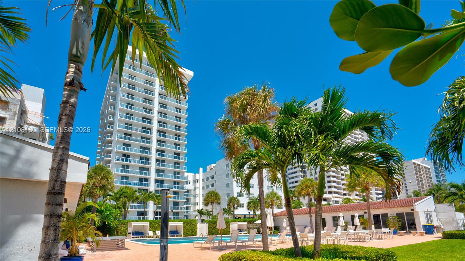 High-rise residential exterior featuring glass-railed balconies, exterior swimming pool, hardscaped patio area, mature palm trees, and manicured landscaping