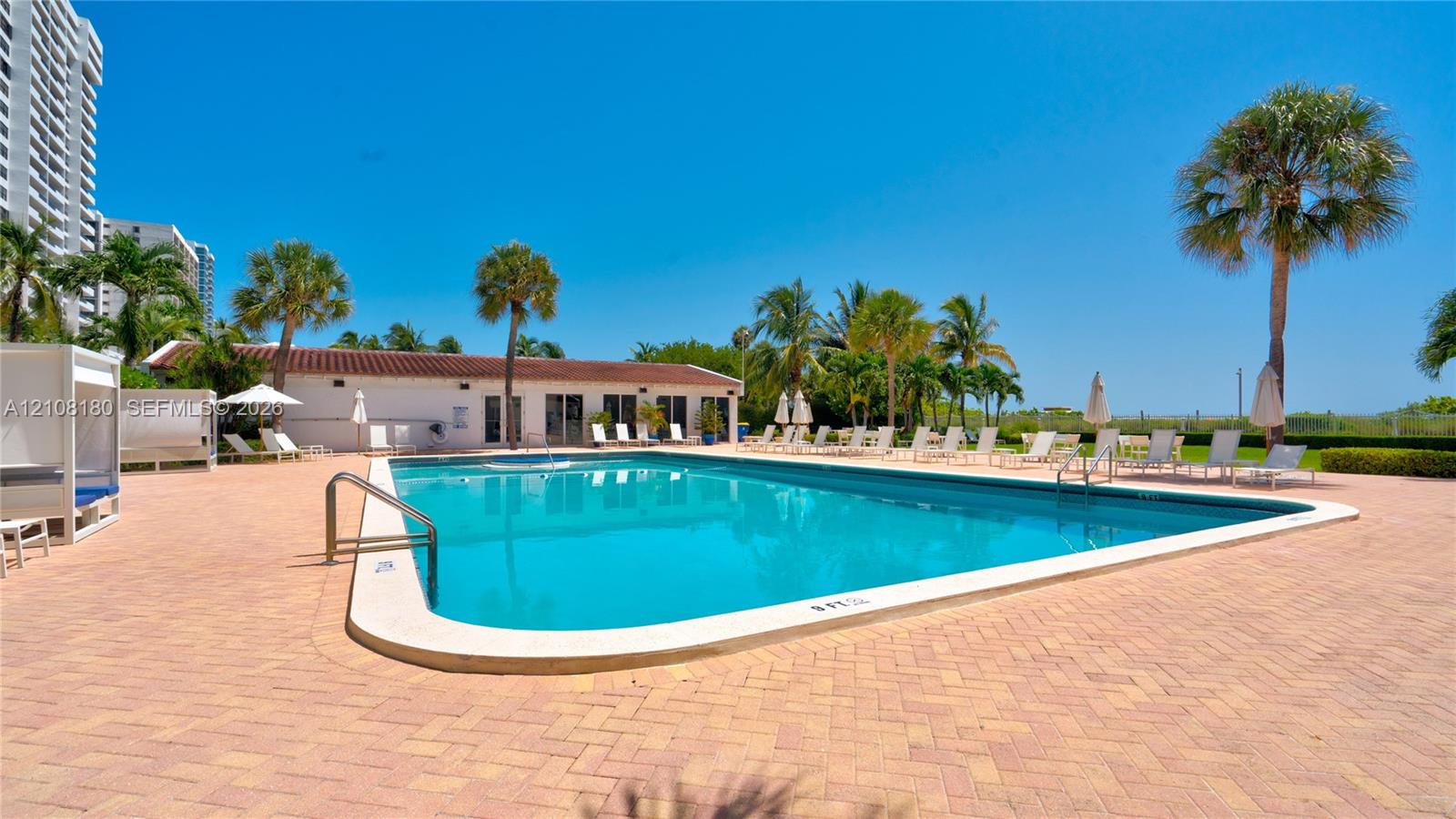 In-ground swimming pool surrounded by a paver deck, palm trees, and a low-profile clubhouse with a clay tile roof