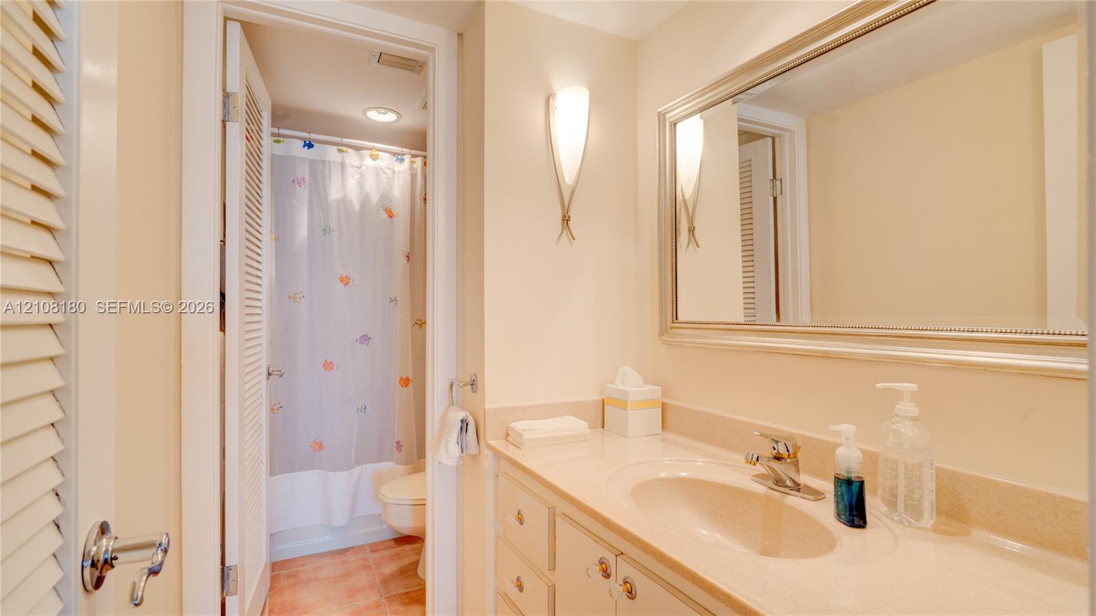 Bathroom vanity with a solid-surface countertop and integrated sink, complemented by a wide framed mirror, wall-mounted sconce lighting, and a separate water closet area featuring a tub-shower combination