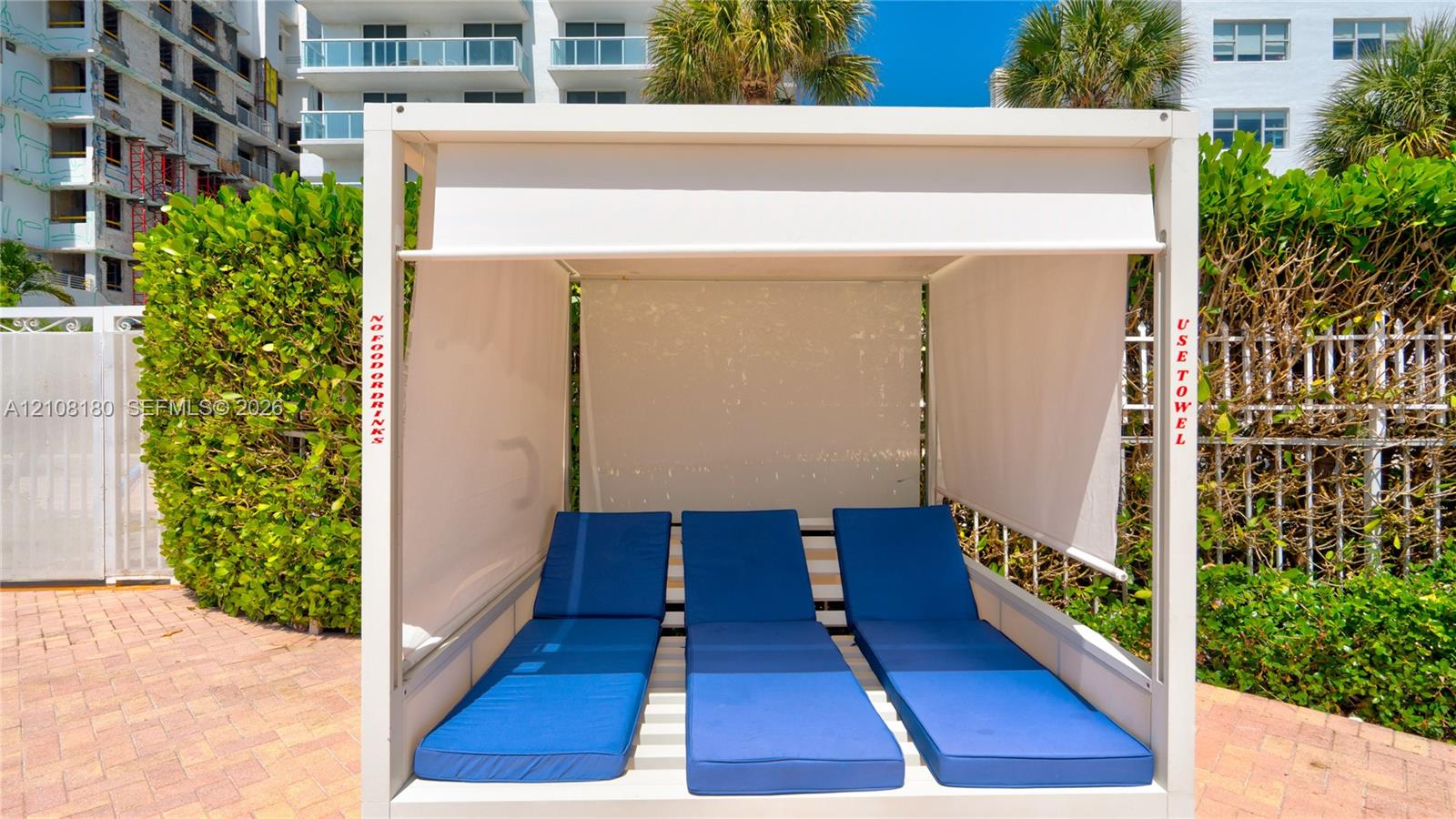 Permanent poolside cabana featuring a white metal frame, integrated retractable fabric shades, and tiered cushioned platforms situated on a brick paver deck
