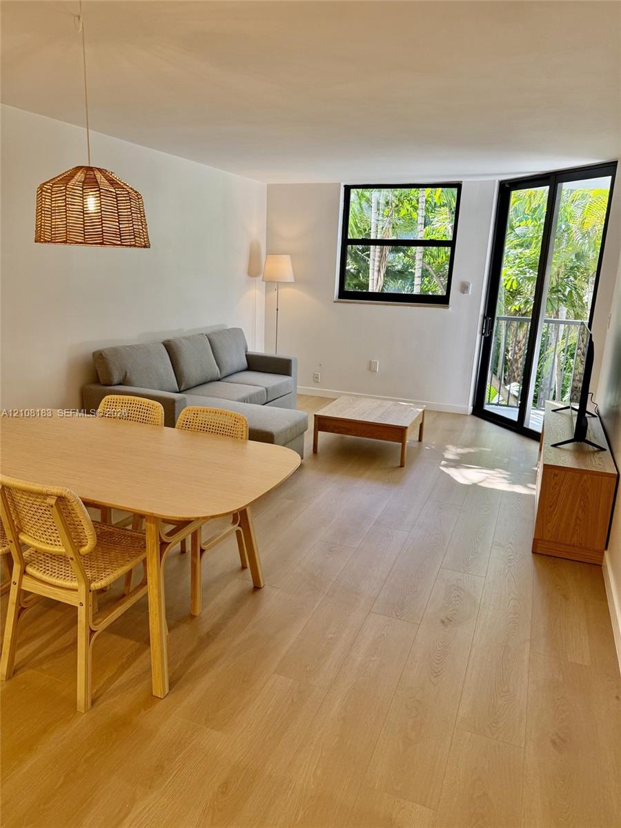 Open-concept living space featuring wood-finish flooring, a woven pendant light fixture, black-framed windows, and glass-paned doors opening to an outdoor balcony