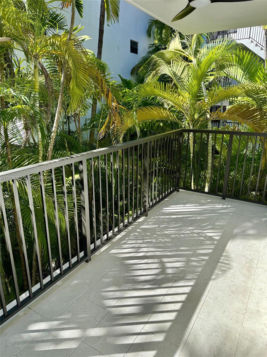 Covered balcony featuring tile flooring, dark metal railing, and an overhead ceiling fan, set against mature palm trees