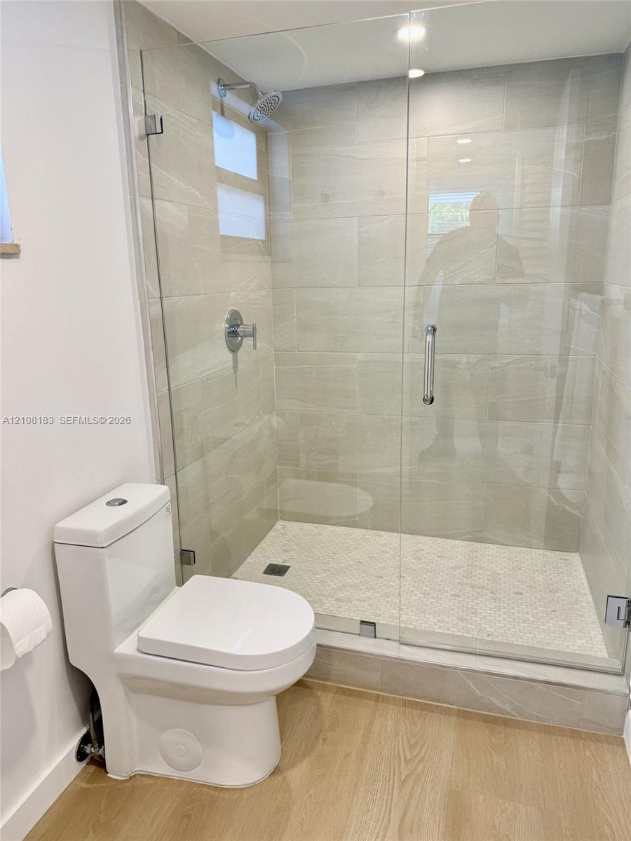 Walk-in shower featuring a glass enclosure, light-toned wall tile, pebble-textured flooring, and a chrome rainfall showerhead, complemented by a dual-flush toilet and wood-finish flooring