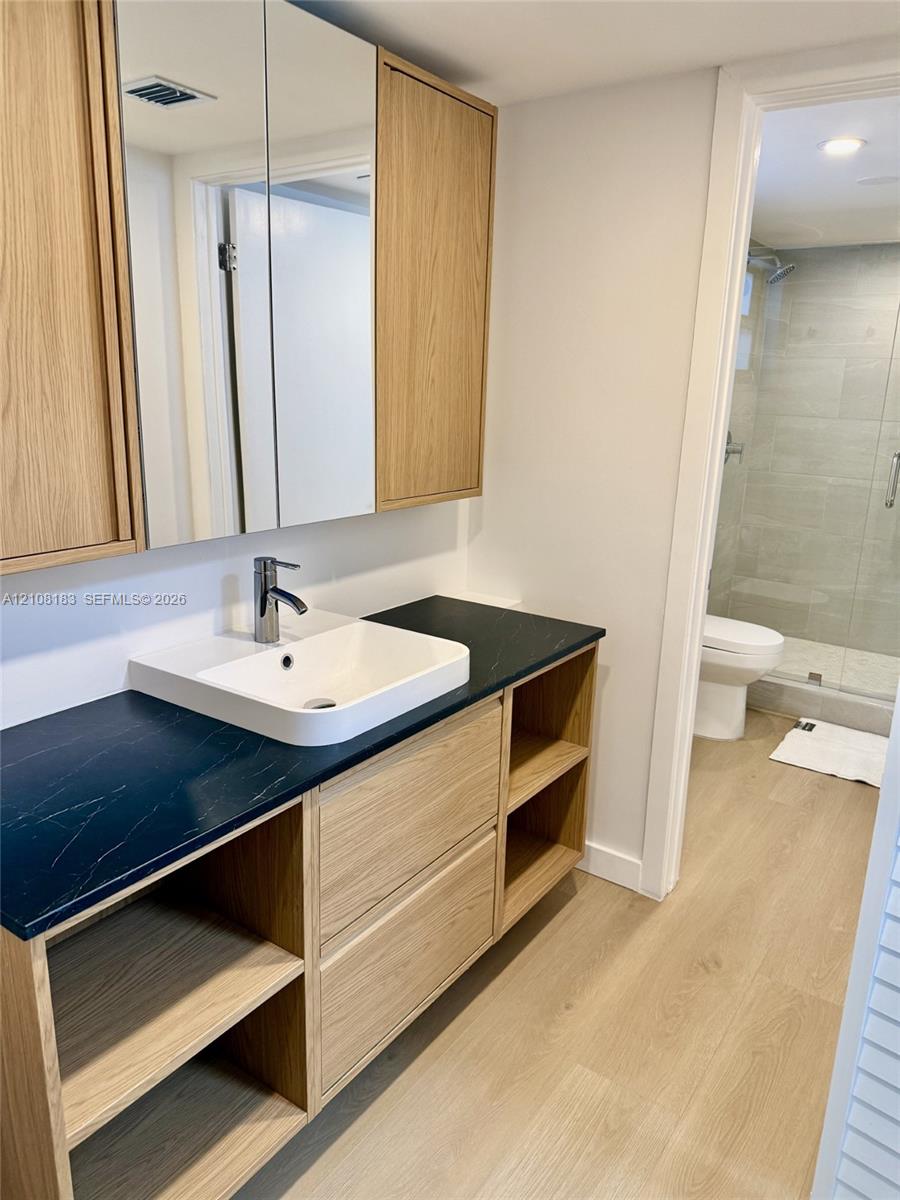 Wood-finish vanity featuring open shelving, a dark countertop, integrated vessel sink, and mirrored medicine cabinets, with wood-finish flooring extending into the tiled shower area