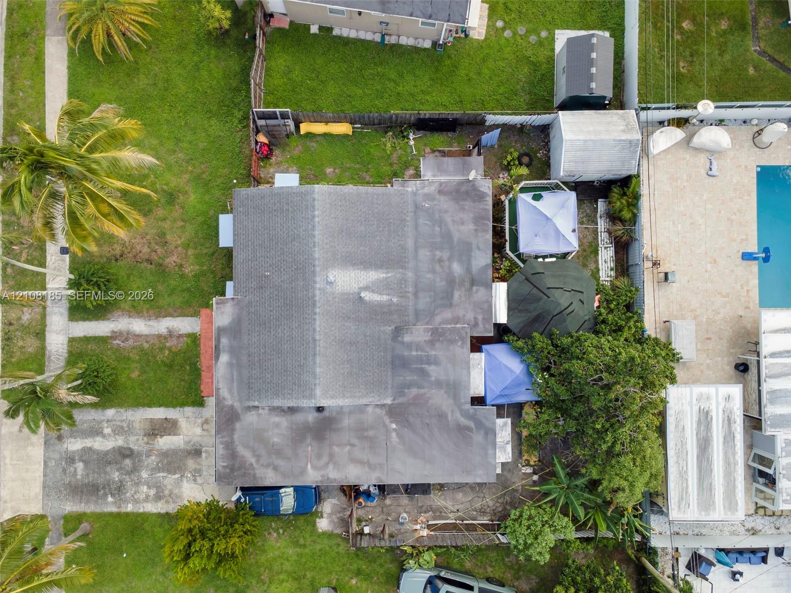 Aerial perspective of a residential lot featuring a multi-section asphalt shingle roof, paved driveway, expansive lawn, and established palm trees