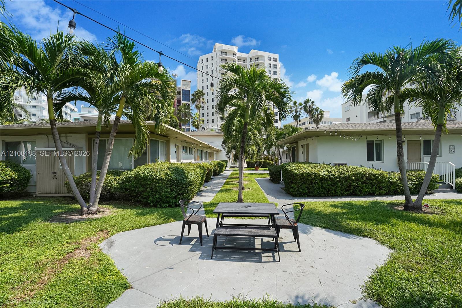 Common area courtyard featuring a concrete patio, manicured hedges, mature palm trees, and overhead string lighting Common area courtyard featuring a concrete patio, manicured hedges, mature palm trees, and overhead string lighting