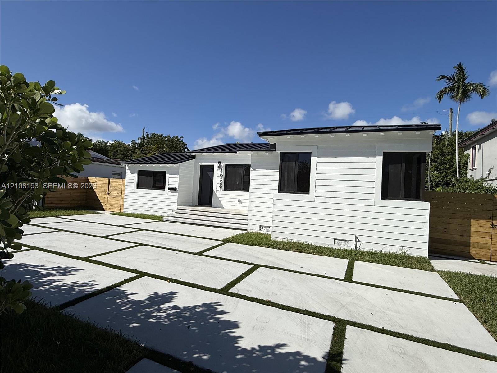 Single-story residence featuring white horizontal siding, a dark metal roof, geometric concrete paver driveway with turf accents, and wood-paneled privacy fencing Single-story residence featuring white horizontal siding, a dark metal roof, geometric concrete paver driveway with turf accents, and wood-paneled privacy fencing