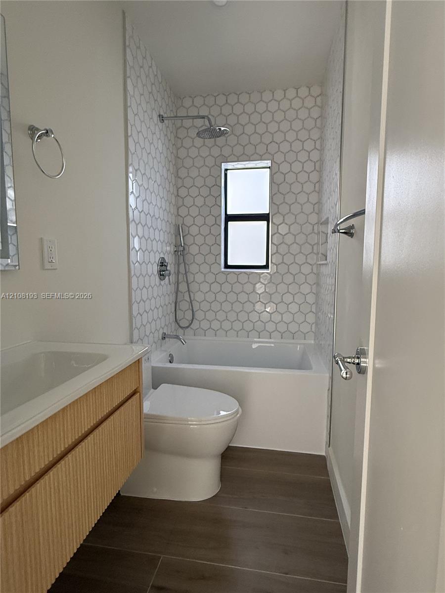 Bathroom featuring a wall-mounted floating vanity with textured wood-finish cabinet, tub-shower combination with floor-to-ceiling hexagonal tile surround, rainfall showerhead, and neutral wood-finish flooring