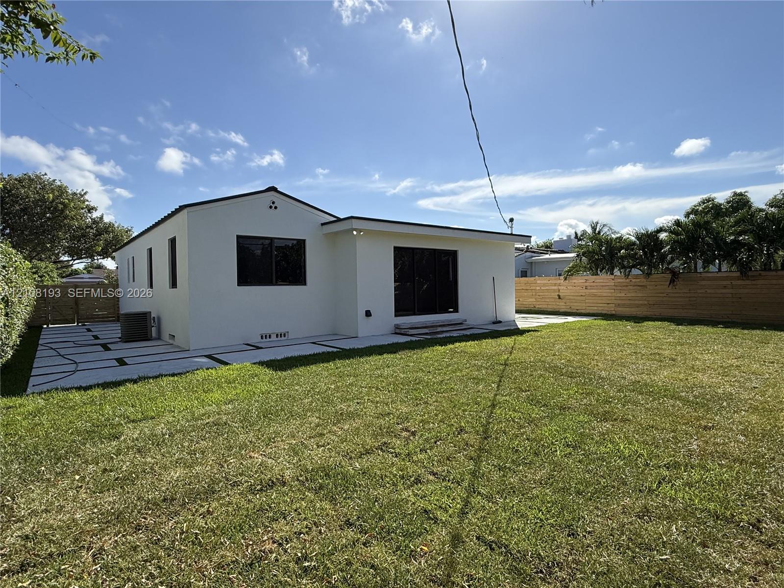 Stucco exterior with a low-pitch roof, expansive grass lawn, large geometric paver patio, and horizontal wood privacy fencing