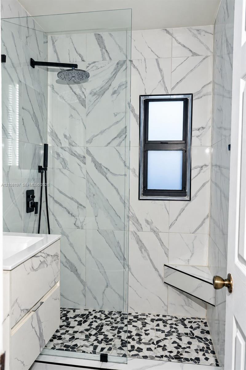 Walk-in shower featuring large-format stone-patterned wall tile, a built-in corner seat, matte black rainfall showerhead with handheld attachment, pebble-texture flooring, and a frameless glass partition
