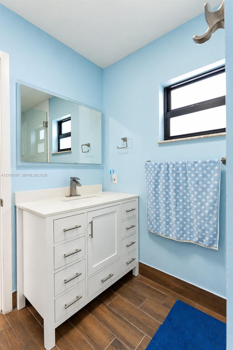 White vanity featuring multiple storage drawers, rectangular basin, and single-lever faucet, complemented by a frameless wall-mounted mirror, wood-finish tile flooring, and metal hardware