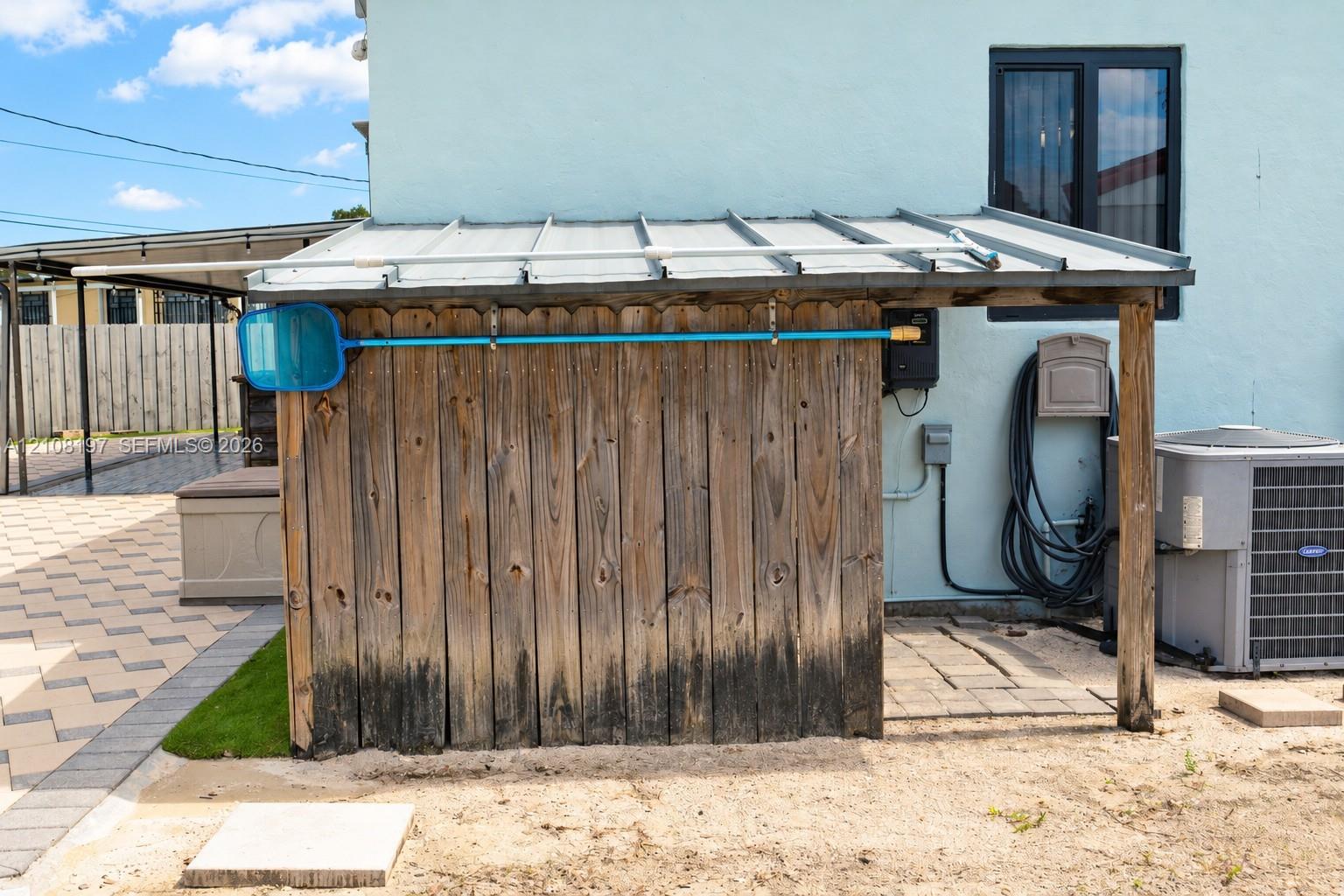 Exterior storage structure with a metal roof and wood vertical planking, featuring an open-sided design, stone paver flooring, and a dedicated central air conditioning unit