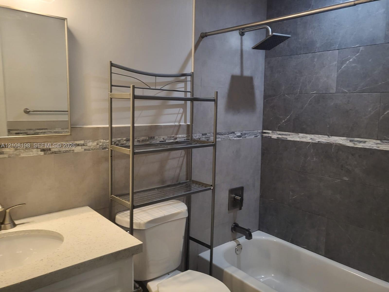 Bathroom featuring a vanity with a light-toned solid surface countertop, rectangular mirror, glass and stone mosaic tile backsplash, and a tub-shower combination with large-format dark tile surround and a rainfall showerhead