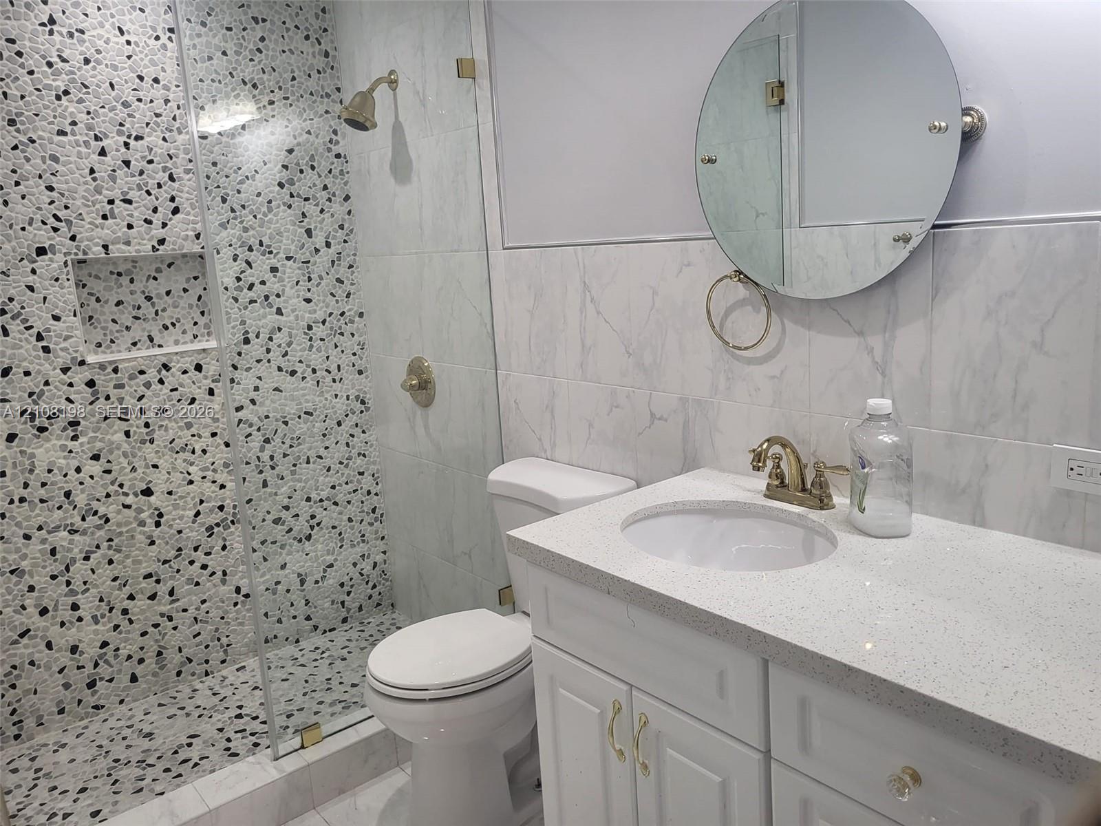 Bathroom featuring a glass-enclosed shower with a pebble-tile accent wall, built-in shower niche, stone-composite vanity top with an integrated sink, and a circular wall-mounted mirror