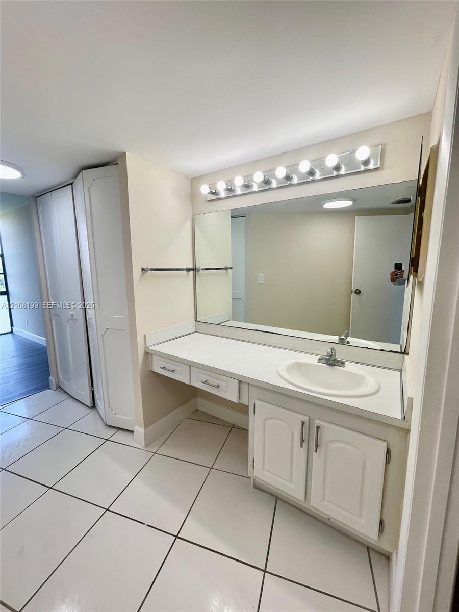 Vanity with white cabinetry, integrated countertop and sink, large rectangular mirror, overhead vanity lighting, and ceramic tile flooring Vanity with white cabinetry, integrated countertop and sink, large rectangular mirror, overhead vanity lighting, and ceramic tile flooring