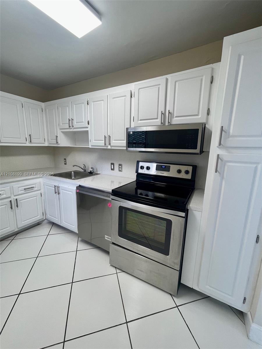 Kitchen featuring white cabinetry, stainless steel appliances, solid-surface countertops, dual-basin stainless steel sink, and large-format tile flooring Kitchen featuring white cabinetry, stainless steel appliances, solid-surface countertops, dual-basin stainless steel sink, and large-format tile flooring