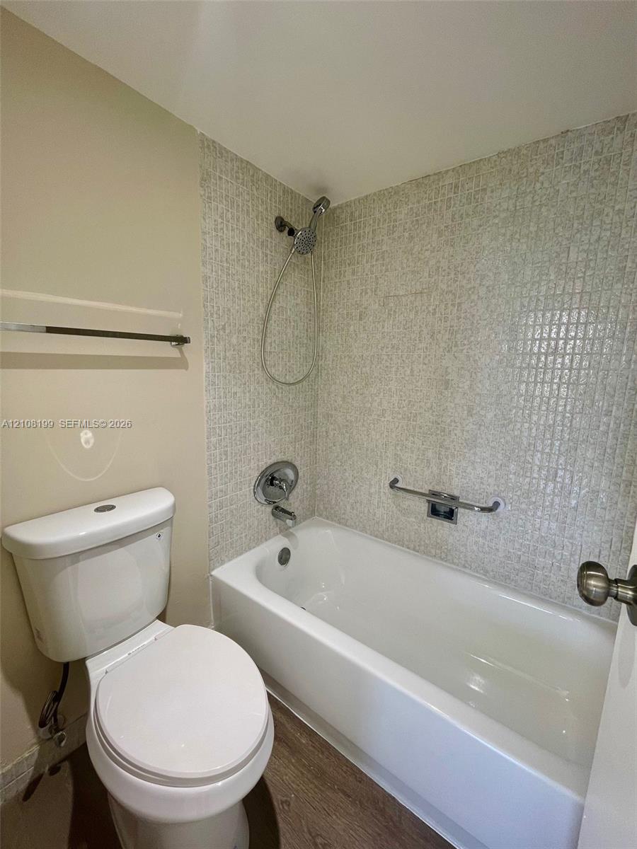 Full bathtub with a handheld shower fixture, glass mosaic tile surround, wood-finish flooring, and wall-mounted metal grab bar