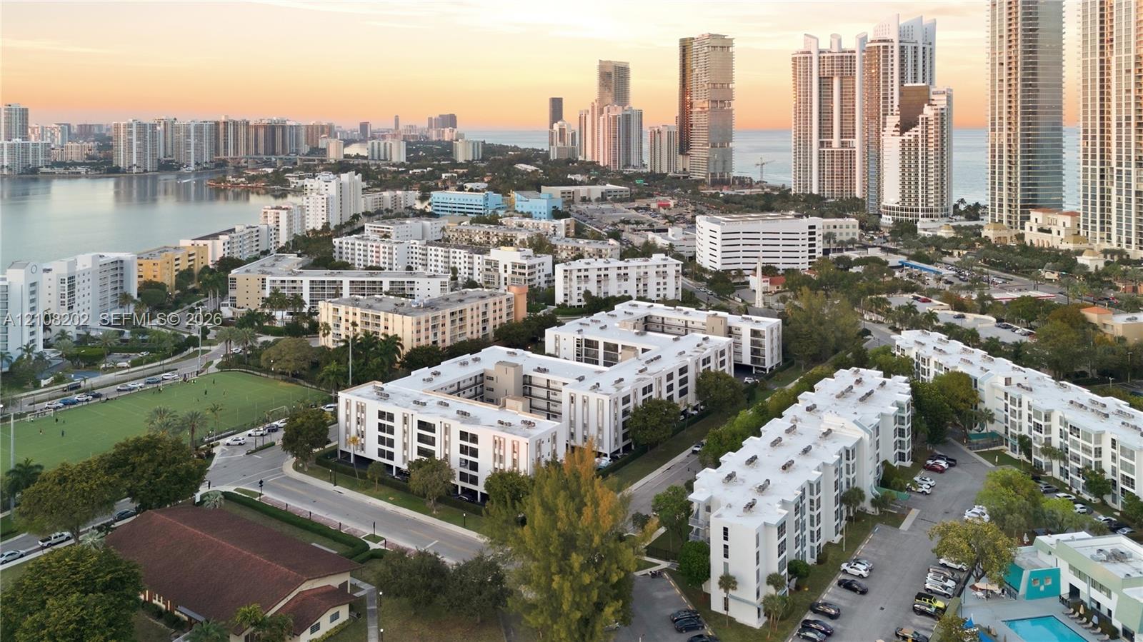 Aerial perspective of the property grounds featuring multi-story residential buildings, paved parking areas, mature landscaping, and proximity to coastal waters and urban skyline