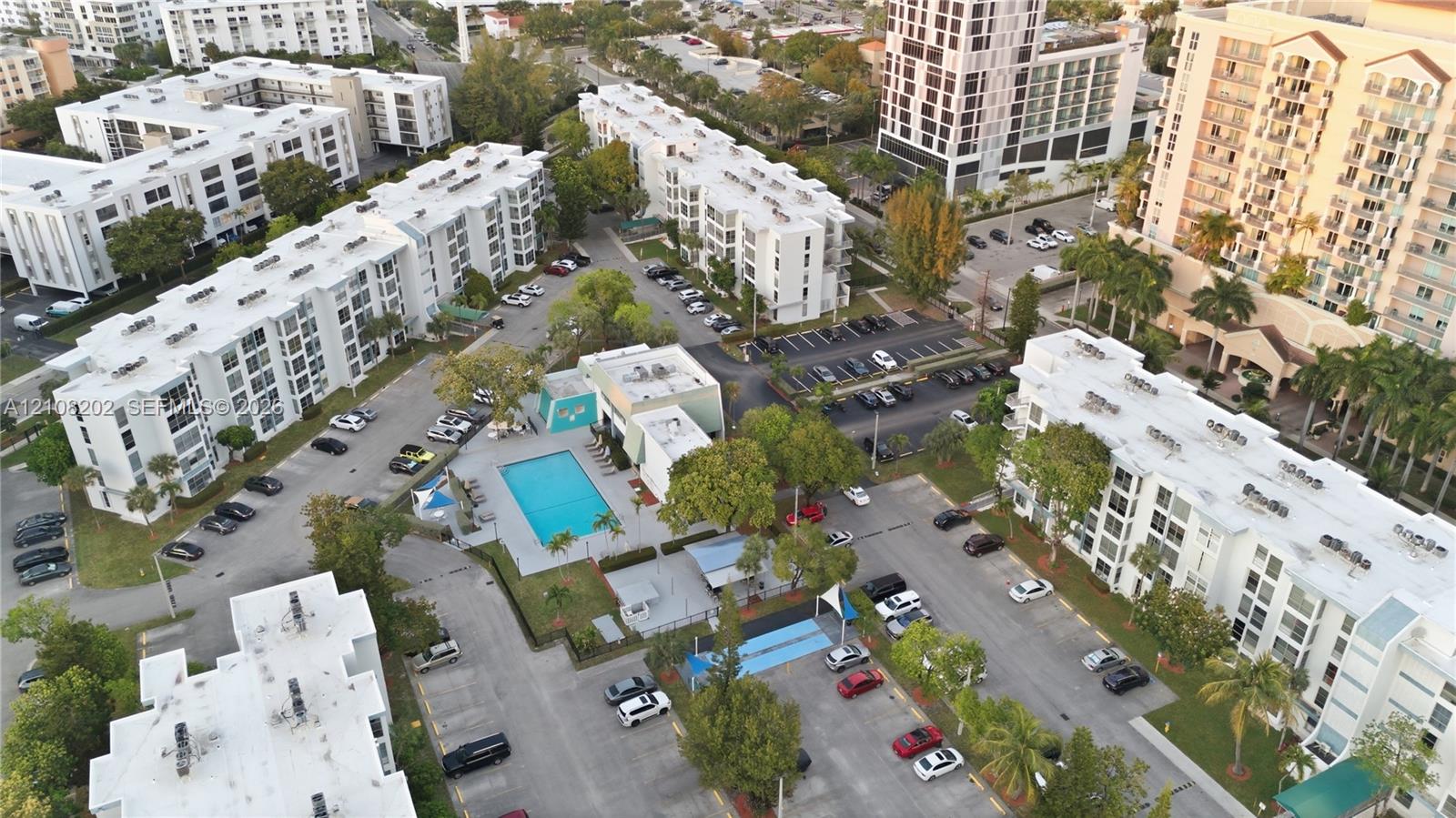 Aerial perspective of a multi-building residential complex featuring a central outdoor swimming pool, paved surface parking, and landscaped grounds with mature trees