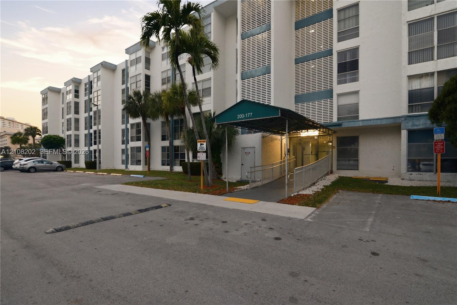 Multi-story exterior featuring a concrete facade, covered entryway with a metal awning, paved surface parking, and mature palm trees Multi-story exterior featuring a concrete facade, covered entryway with a metal awning, paved surface parking, and mature palm trees